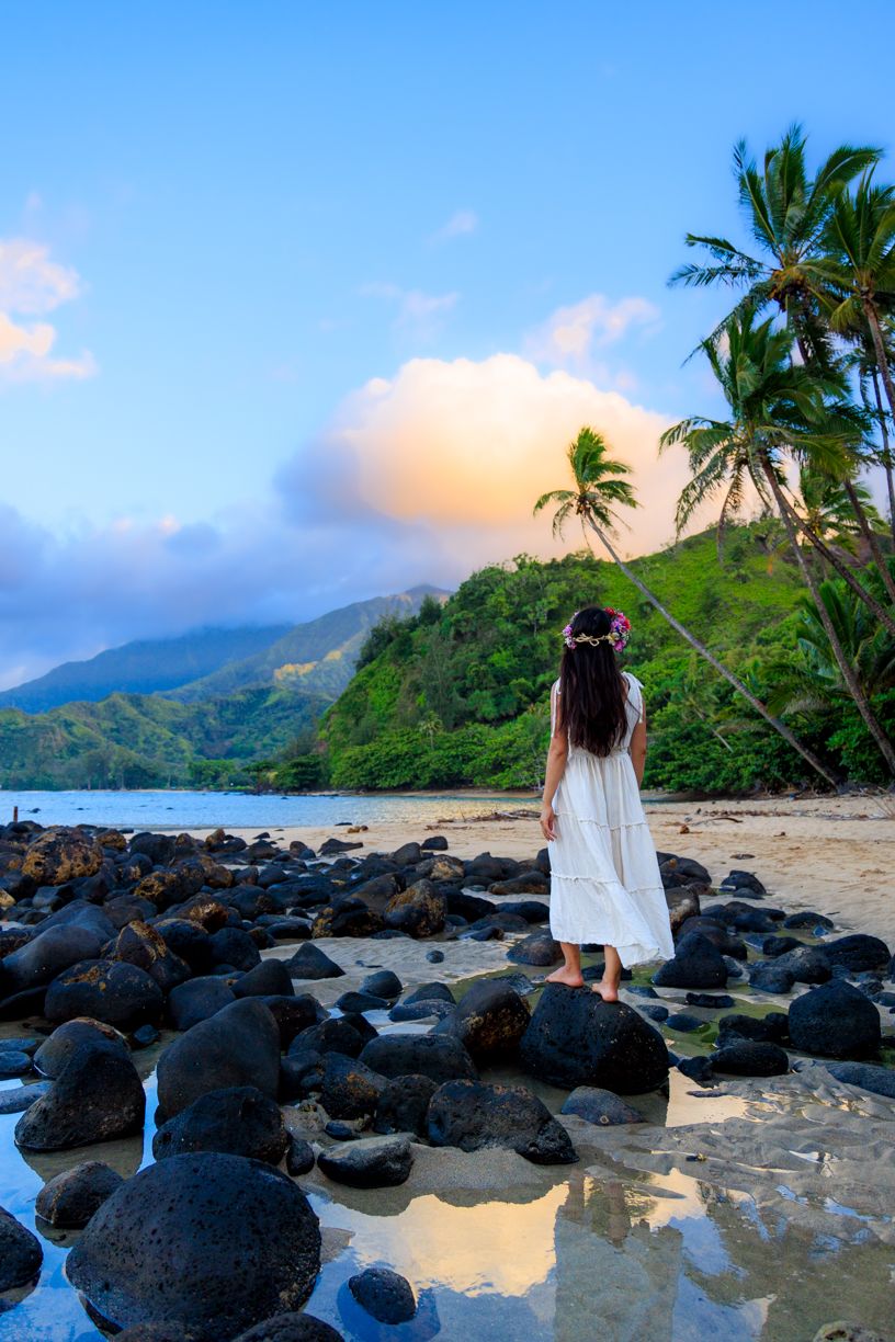 Person in white dress stands on rocky beach with palm trees and mountains in Hawaii at sunset.