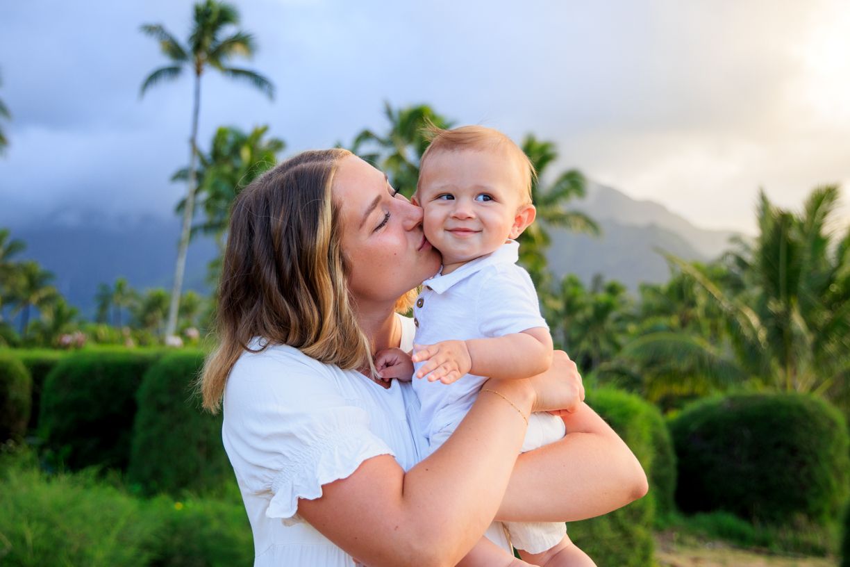Mother kissing baby in tropical setting with palm trees and mountains in background.
