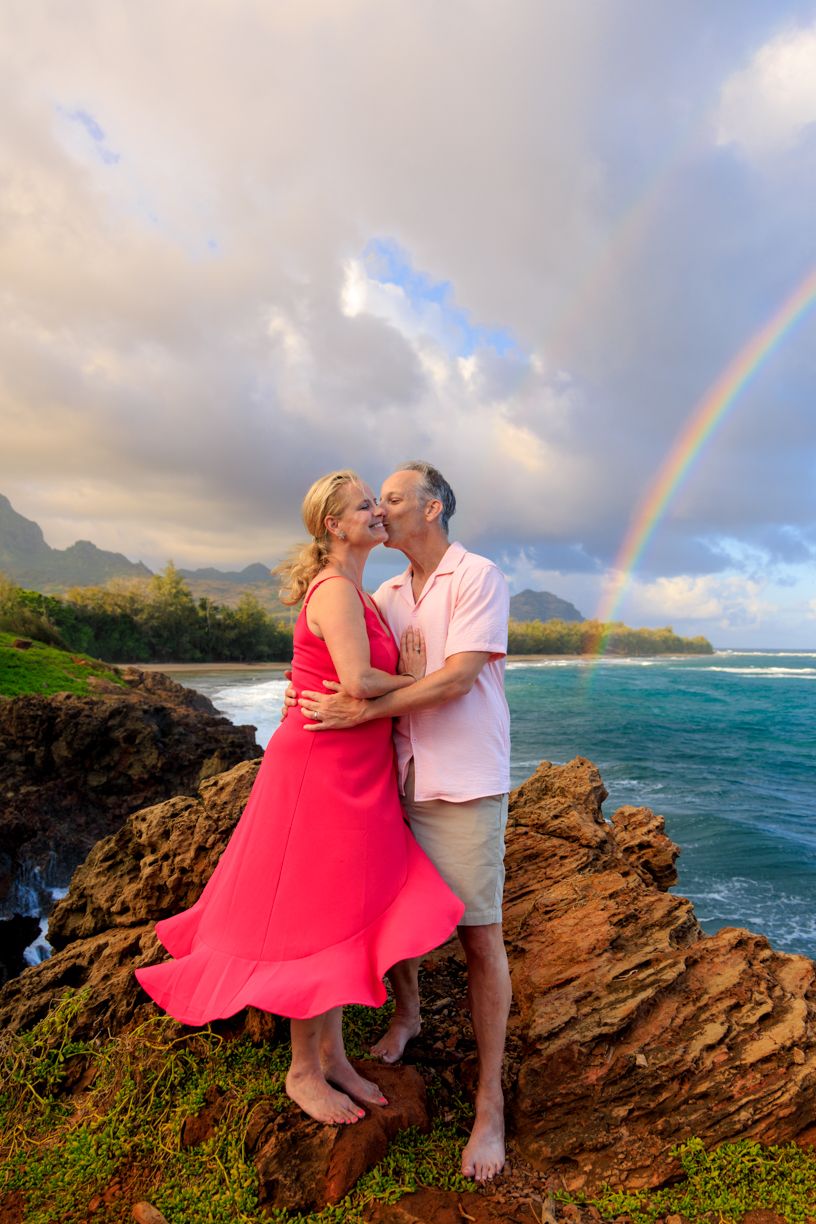 A couple embraces on rocky cliffs in Hawaii with a rainbow and turquoise ocean waters in the background.