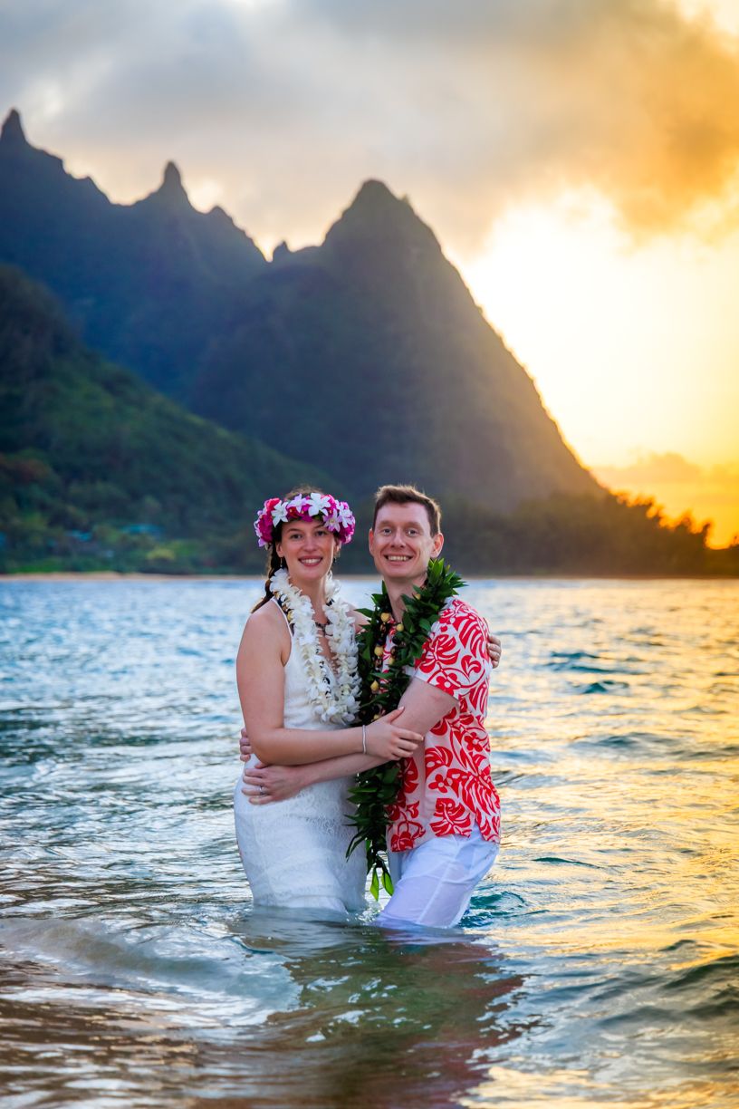 A couple enjoys a playful moment in the ocean during sunset with mountain silhouettes in the background.