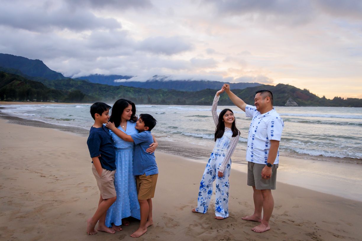 A group gathers on a scenic Hawaiian beach at sunset with mountains and ocean waves in the background.