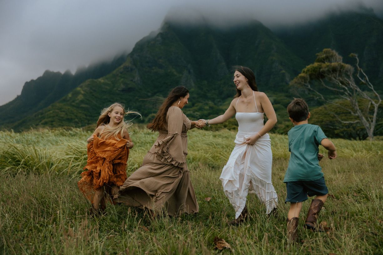 Women and children dance joyfully in a lush Hawaiian field with dramatic misty mountains in the background.