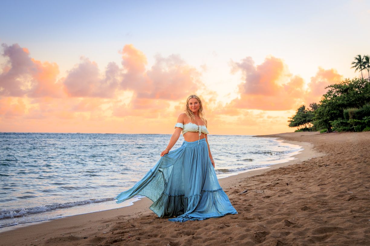 Person in blue dress sitting on beach at golden sunset in Hawaii.