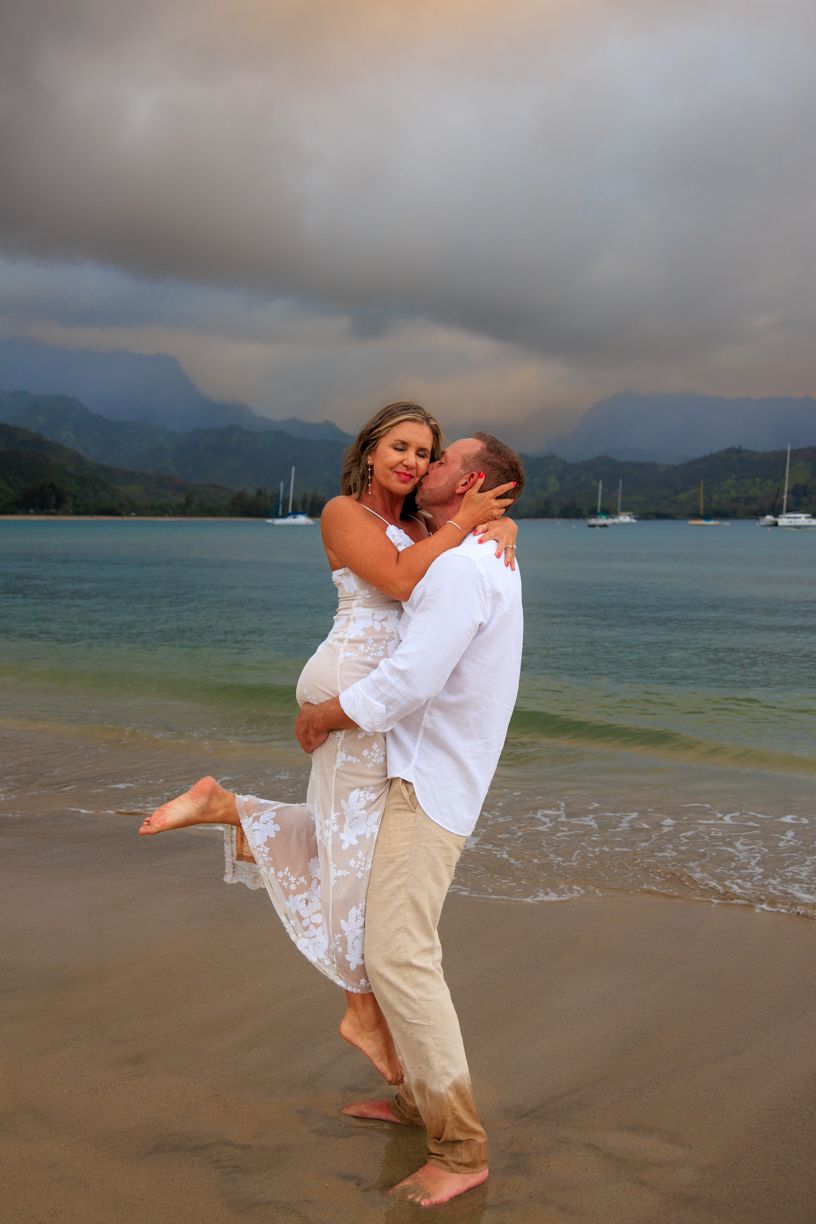 A romantic beach wedding photo with mountains and boats in the background.