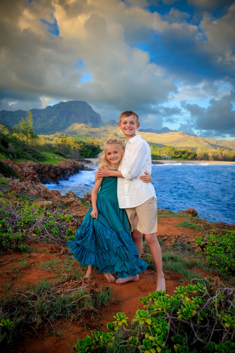 Couple in teal dress and white shirt photographed on rocky coastline with mountains and ocean view.