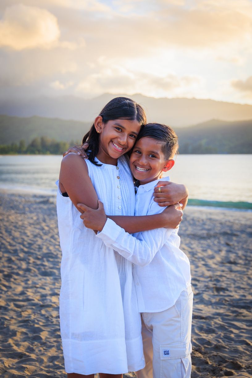 Children in white outfits sharing joyful embraces on a beach at sunset.