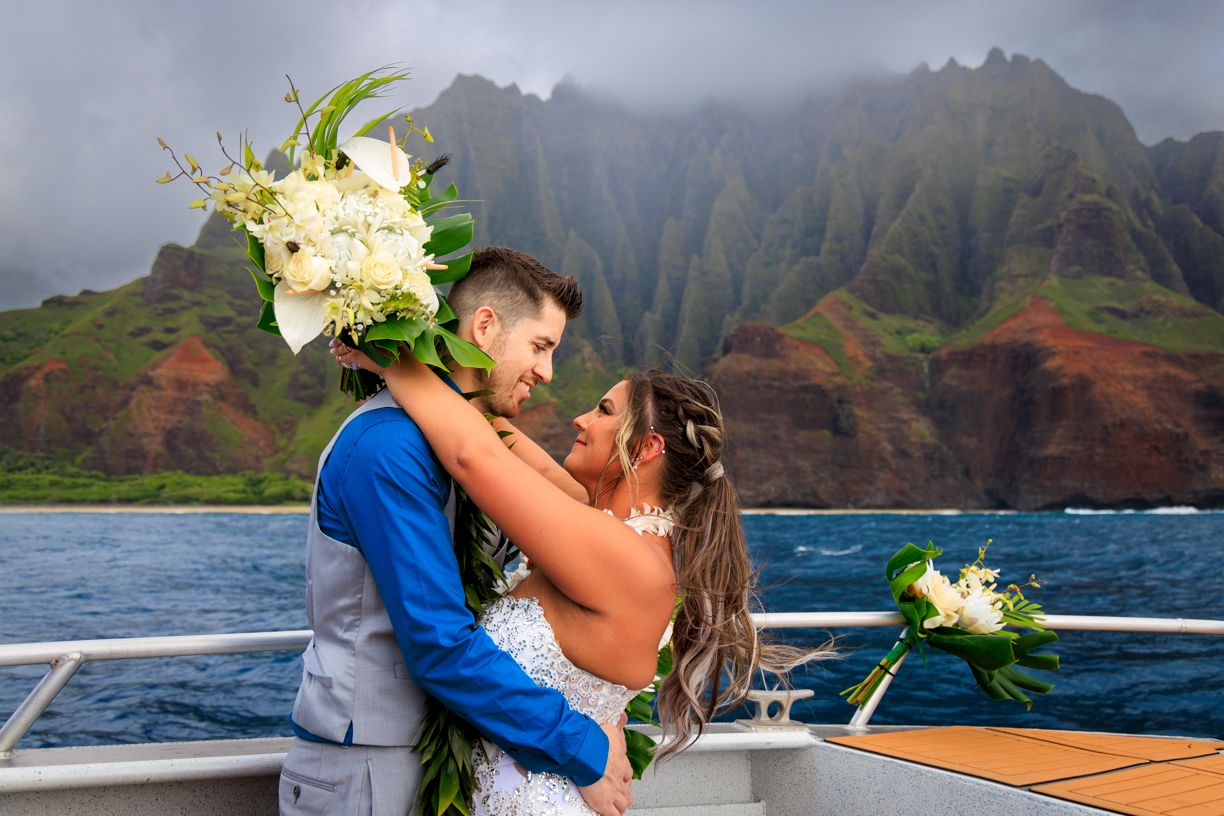 A couple embraces on a boat with the dramatic Na Pali Coast of Kauai in the background.