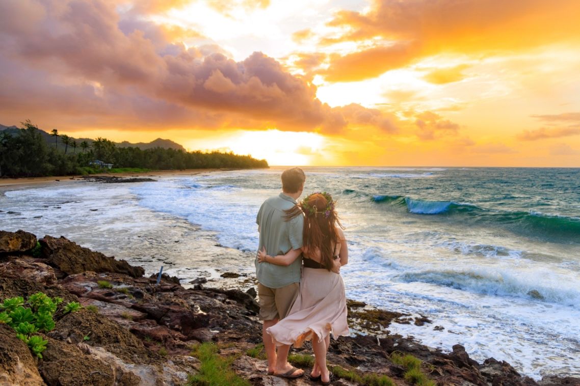 Couple watches dramatic orange sunset over Hawaiian beach coastline.