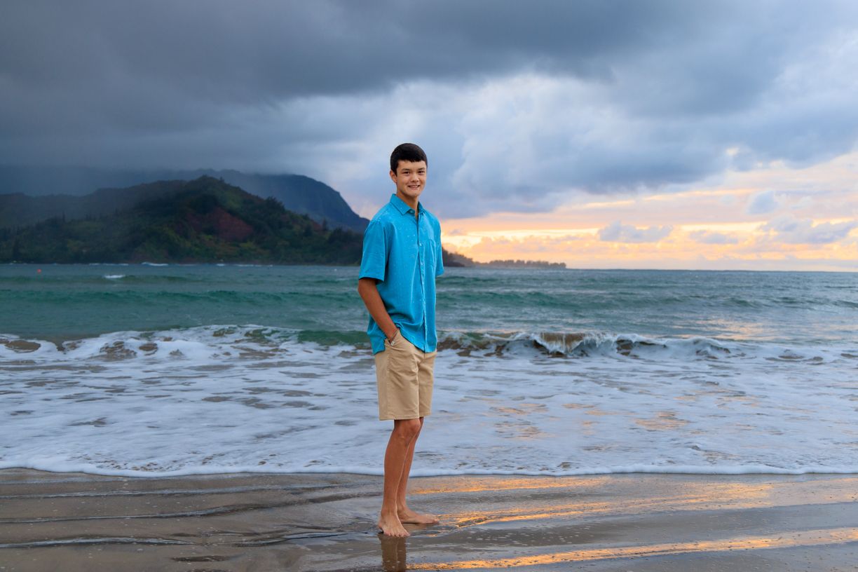 Man in blue shirt and khaki shorts standing on beach at sunset with mountains in distance.