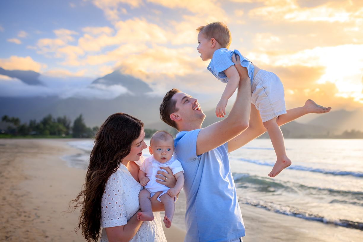 Family of four enjoying a beautiful beach sunset, parents lifting babies with mountains and golden sky in background.