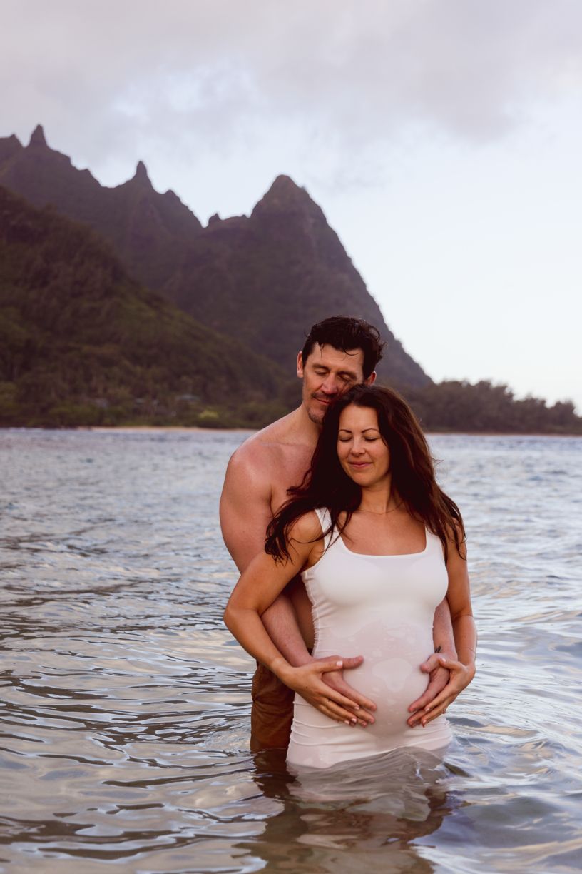 A maternity photoshoot in the ocean captures a couple against mountainous Hawaiian scenery.