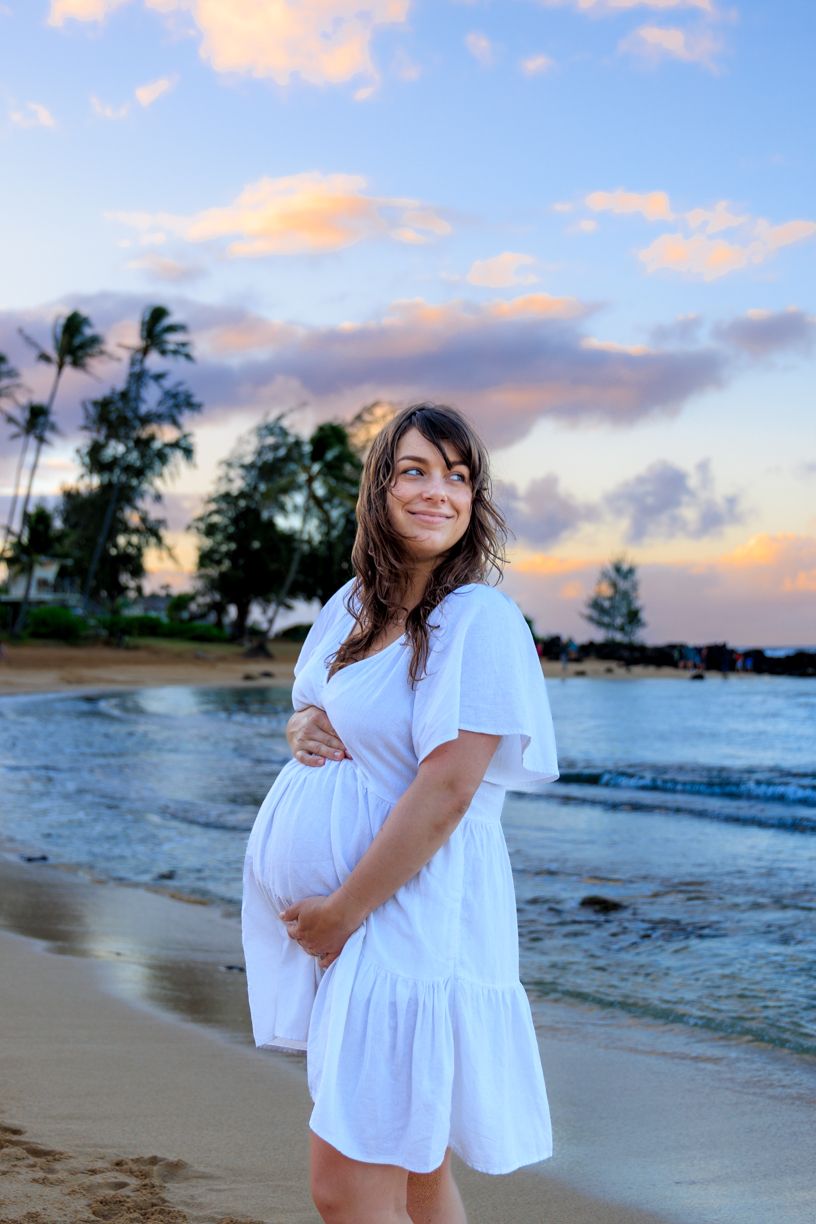 Beachside maternity portraits in white dresses with palm trees and sunset clouds.