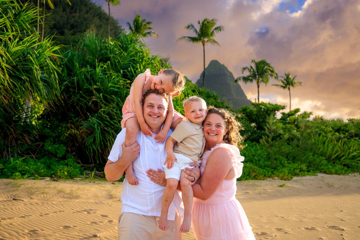 A family shares a joyful moment on a Hawaiian beach with mountains and palm trees at sunset.