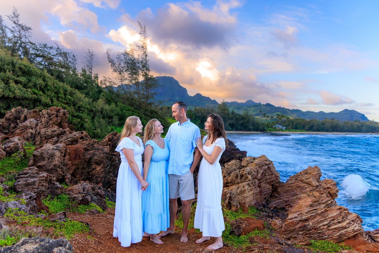 A group poses on rocky coastline with dramatic mountain and ocean views at sunset.