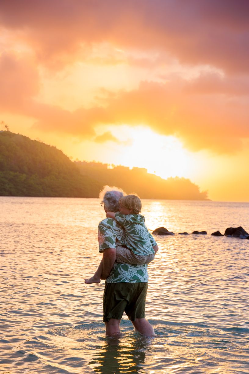 Elderly couple embracing while standing in shallow ocean water during a vibrant golden sunset near a tropical hillside.