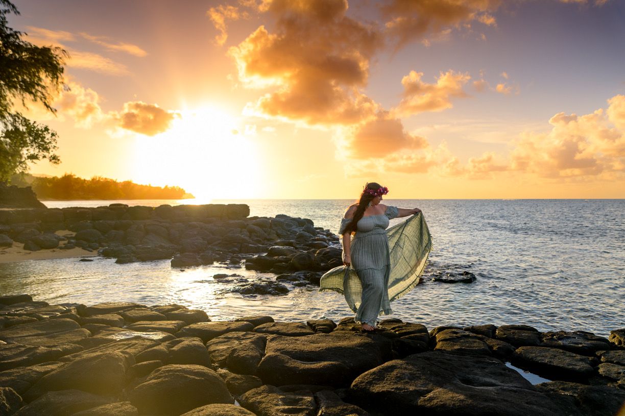 A flowing dress catches the golden light at sunset along a rocky Hawaiian coastline.