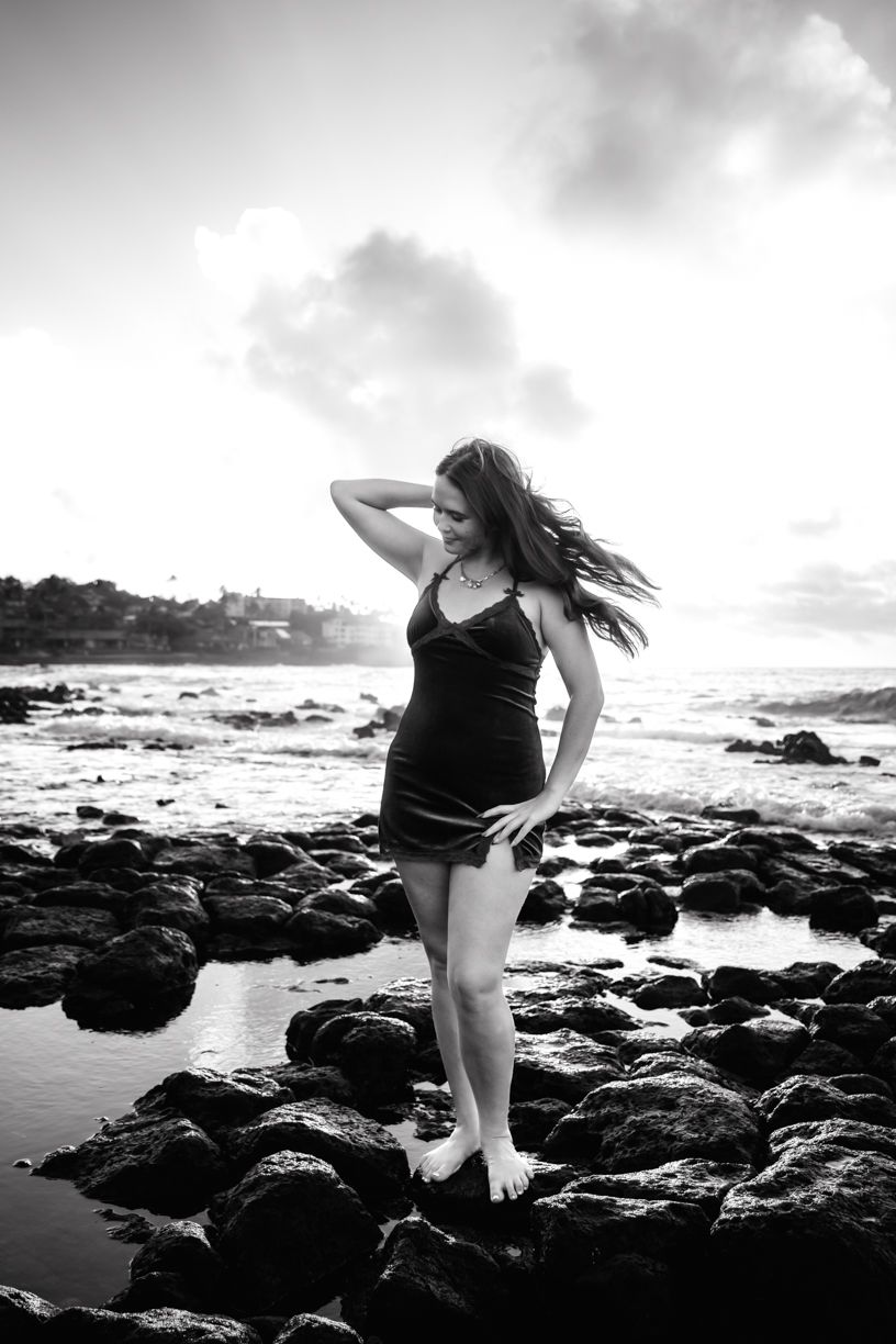 Black and white photo of person posing on rocky shoreline with dramatic sky and ocean waves.