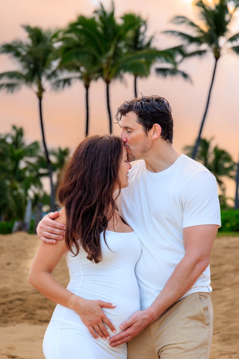 A romantic couple in white outfits embrace on a tropical beach with palm trees at sunset.