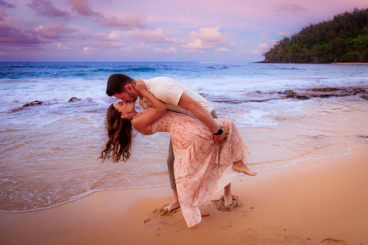 Romantic dip pose on sandy beach with crashing waves and pink sunset sky.