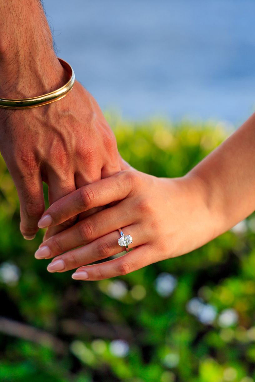 Close-up of hands showing off a sparkling engagement ring against a tropical backdrop.