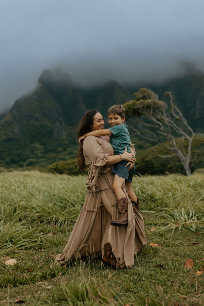 A mother in a tan dress holds her young son in a lush Hawaiian valley with misty mountains behind them.