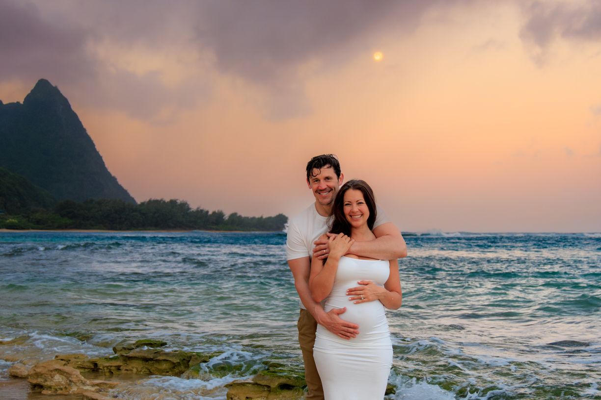A couple embraces on a tropical beach with dramatic mountain peaks and ocean waves during golden hour.