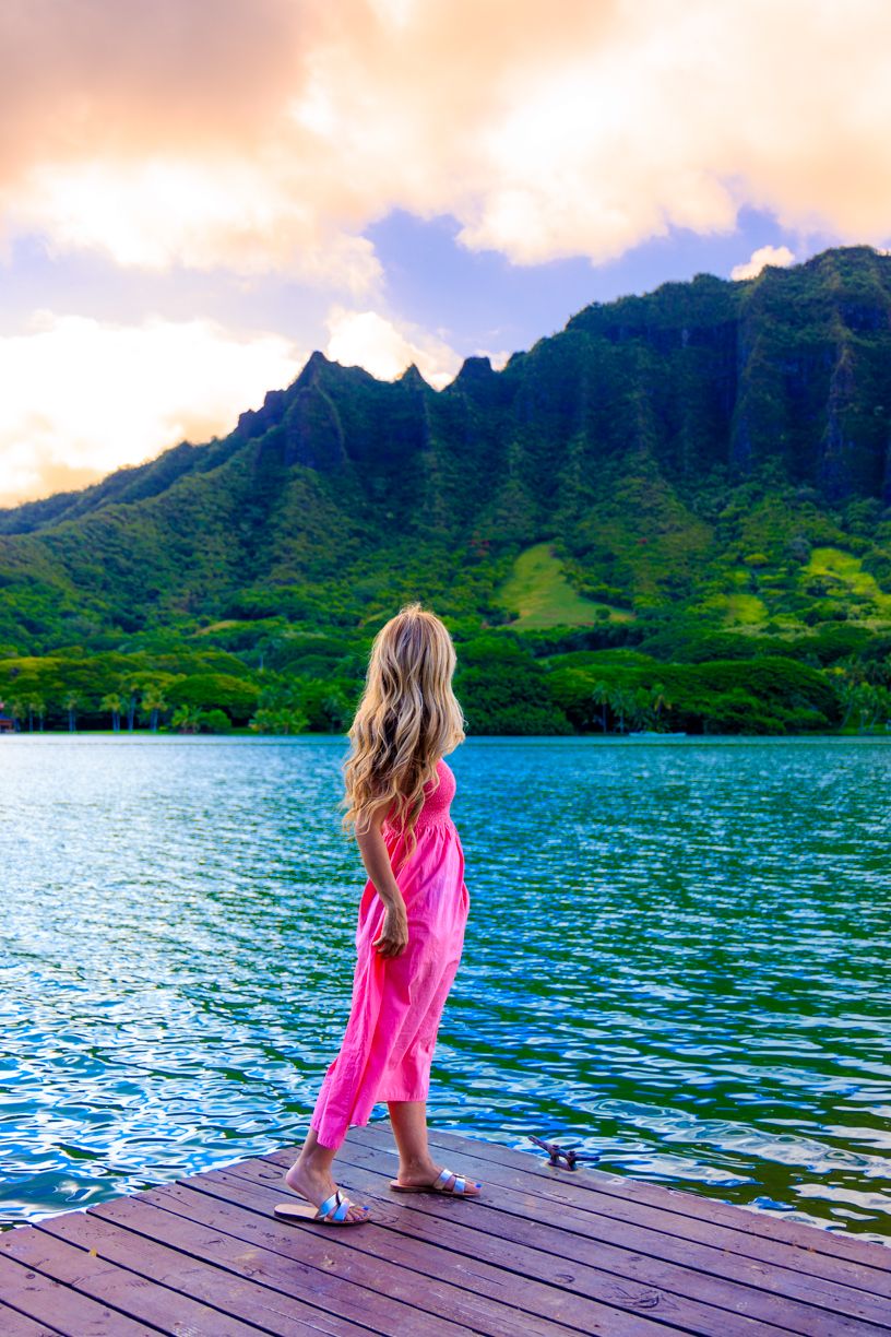 Person in bright pink dress stands on dock overlooking mountain-ringed tropical lake.