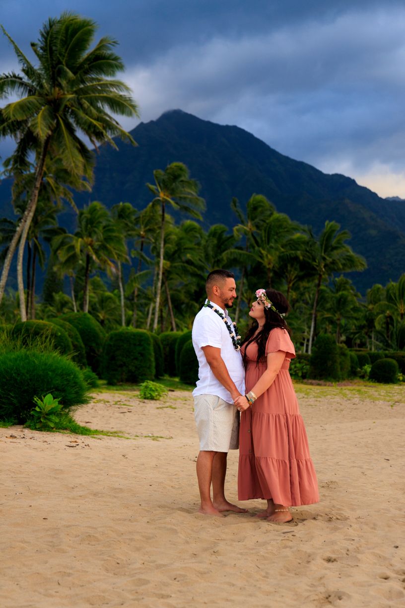 A couple shares an intimate moment on a sandy beach with mountains and palm trees behind them.