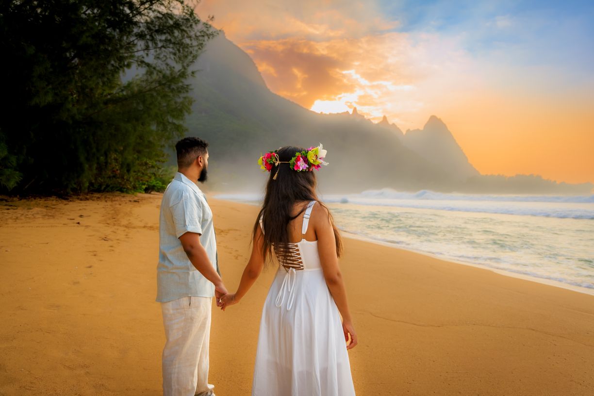 Couple walking along sandy beach at golden hour with dramatic sunset sky.