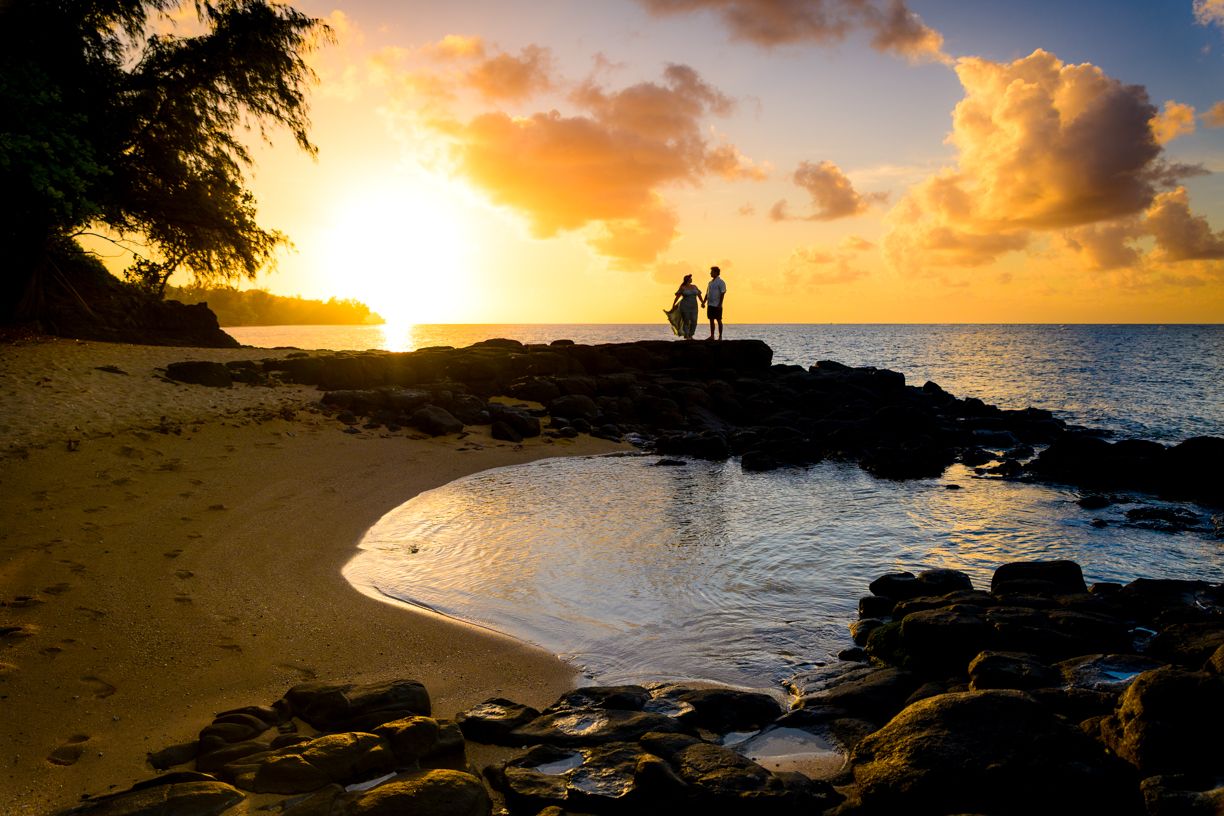 Two silhouettes stand on rocky shore during golden sunset in Hawaii.