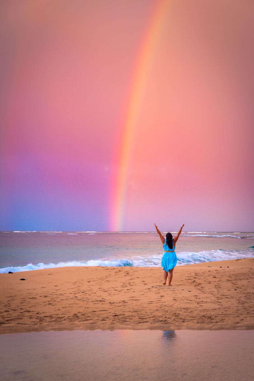 Vibrant rainbow appears over sandy beach during pink sunset.