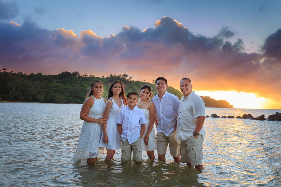 Family of six posing on a tropical beach at sunset with vibrant orange and purple skies reflecting on calm waters.