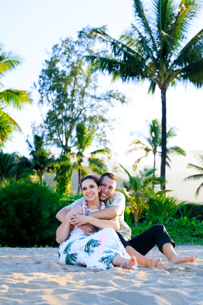 Two people pose playfully on a sandy beach with palm trees during a sunset photoshoot.