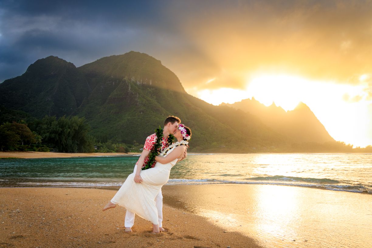 Couple in wedding attire dancing on beach at sunset with mountains in background.