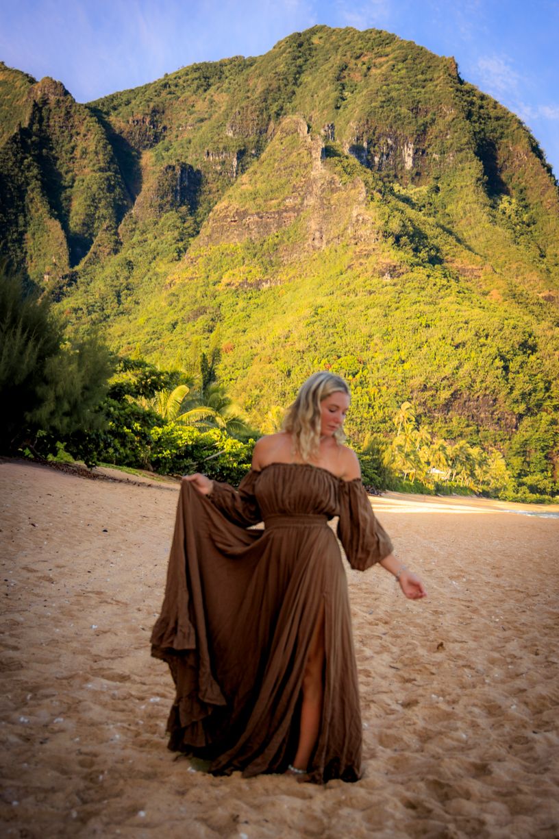 Flowing brown dress photographed against dramatic mountain landscape on sandy beach at sunset.