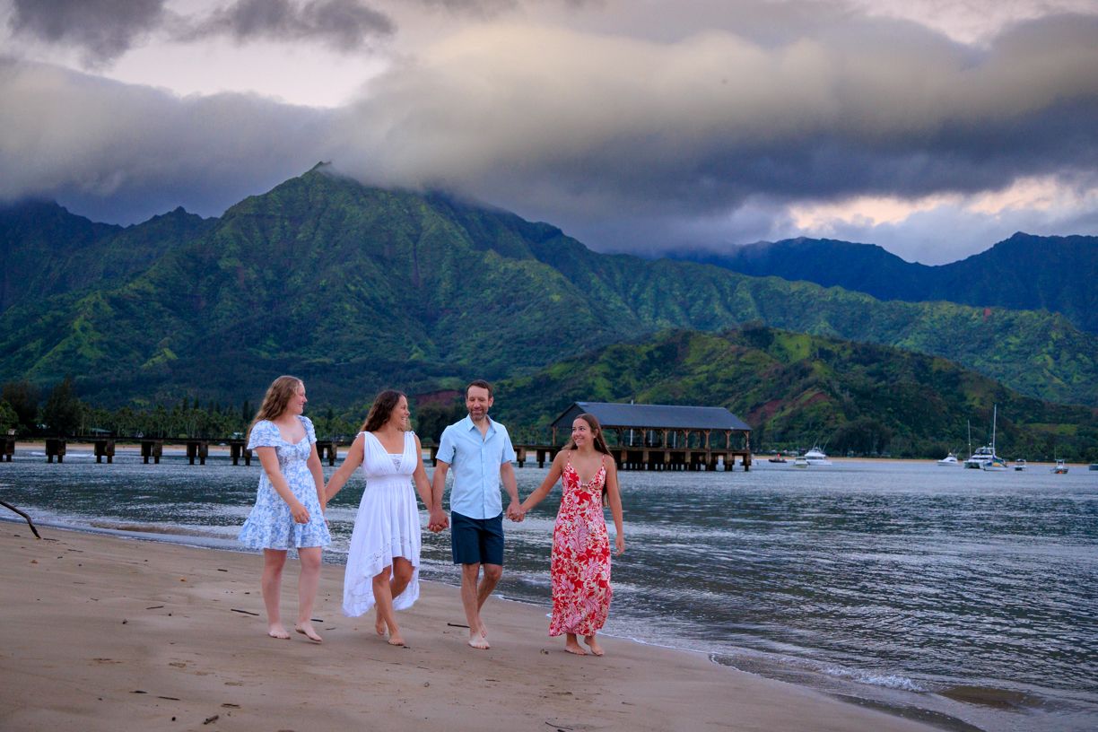 Family group walking along a scenic beach with mountains and pier in the background at dusk.