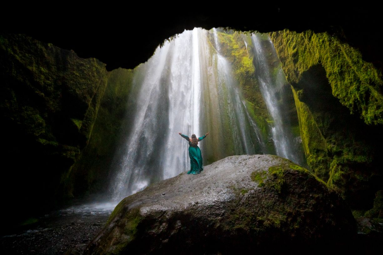 A figure stands on a rock beneath a dramatic waterfall in a moss-covered cave.