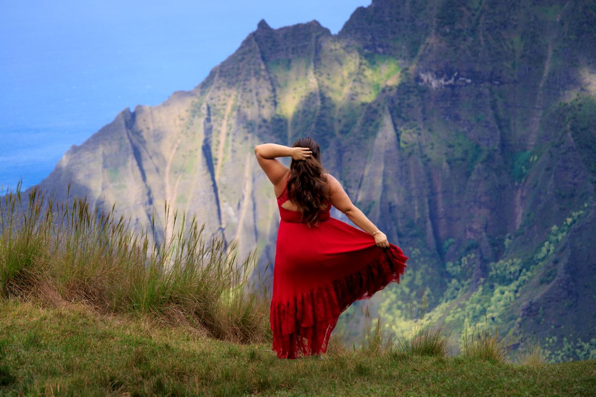 A red dress flows in the wind against dramatic mountain cliffs in Hawaii.