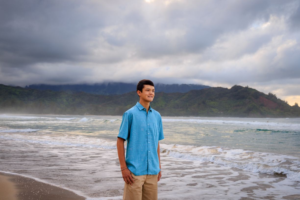 Man in blue shirt standing on beach with mountains and cloudy sky in background.