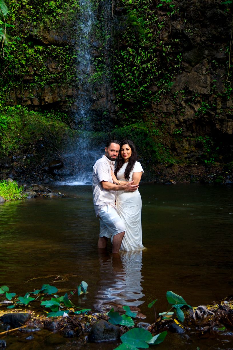 A couple stands together in a serene pool beneath a cascading waterfall surrounded by lush greenery.