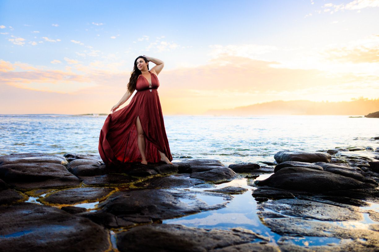 Person in burgundy dress poses on rocky coastal outcrop during golden hour.