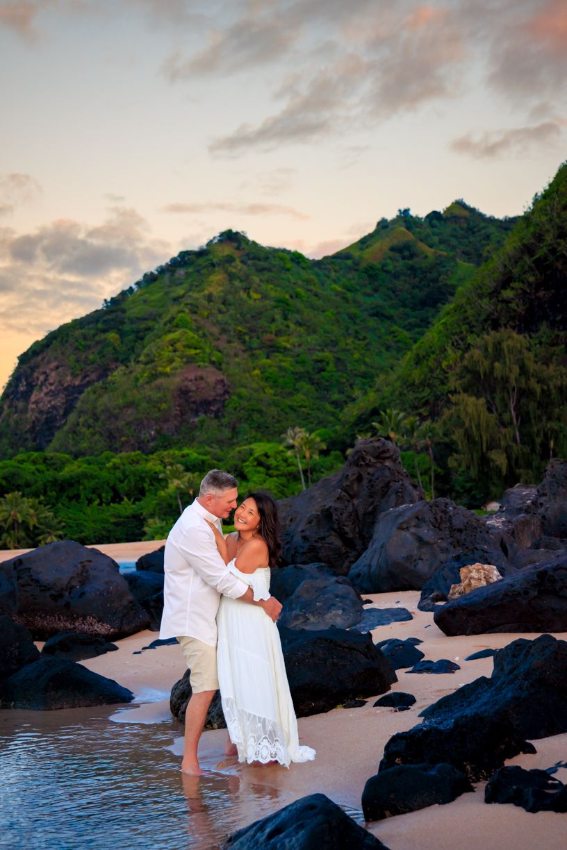 Beach wedding photos at sunset with volcanic rocks and tropical mountains in background.