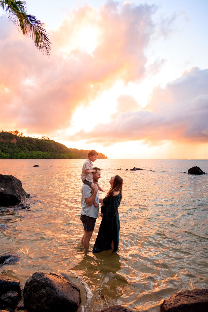 Family standing in ocean water at sunset, with child on father's shoulders, tropical beach and colorful sky backdrop.