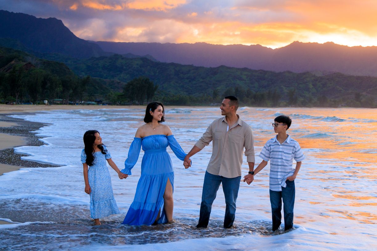 Family of four holding hands on a Hawaiian beach at sunset with mountains and vibrant orange sky.