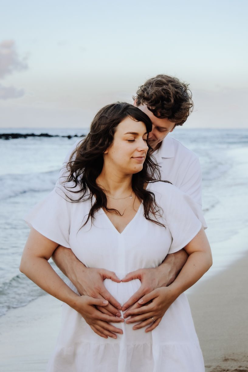 A couple creates a heart shape with their hands over a baby bump during a beach photoshoot.