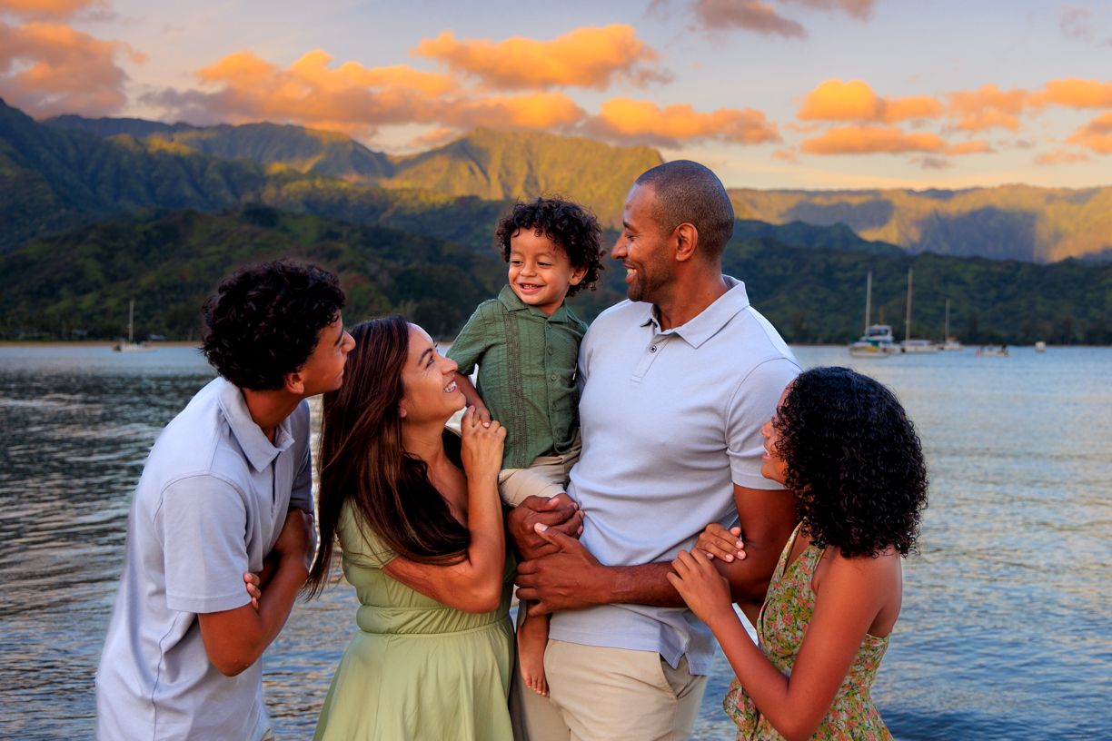 A loving family embraces during a golden sunset on a Hawaiian beach with mountains in the background.