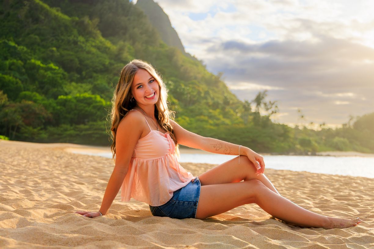 Woman in pink top and shorts sitting on beach with mountains and ocean at sunset.
