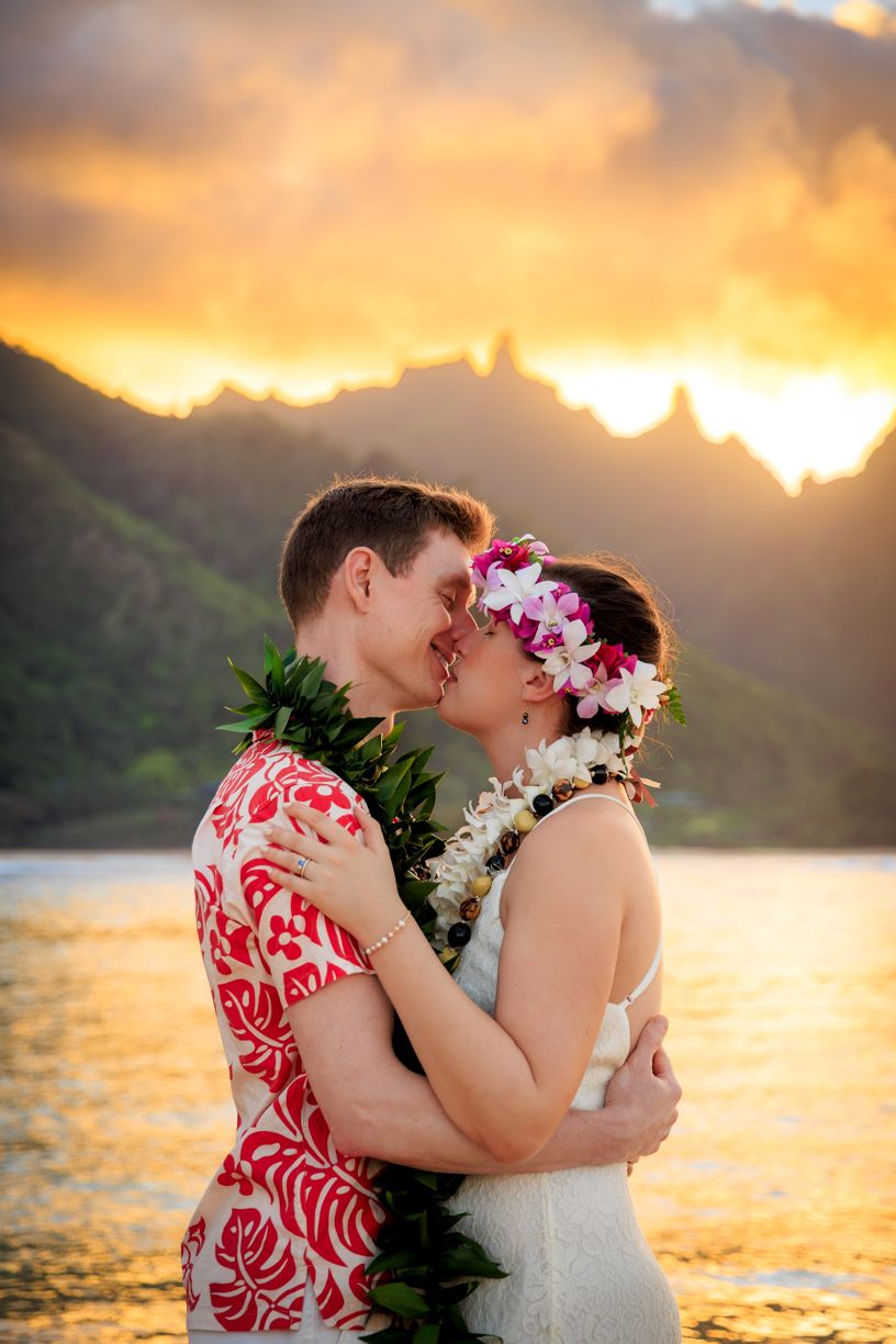 Couple in traditional Hawaiian wedding attire embracing at sunset by the ocean.