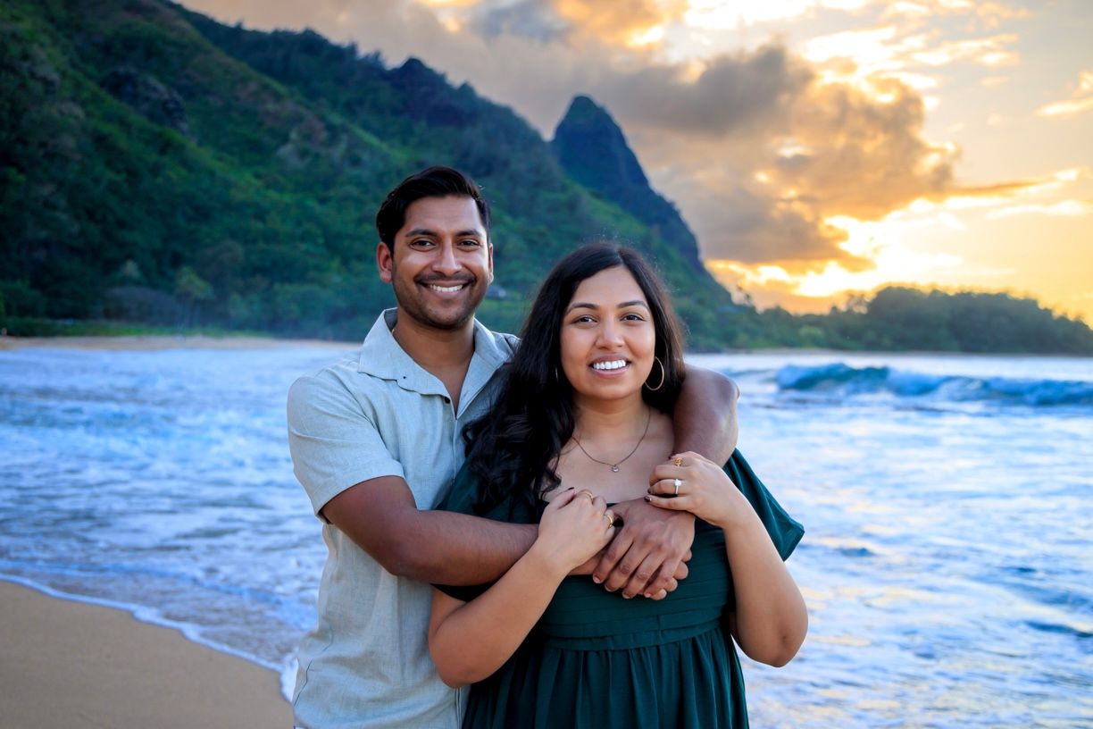 A romantic couple embraces on a Hawaiian beach at sunset with mountains in the background.