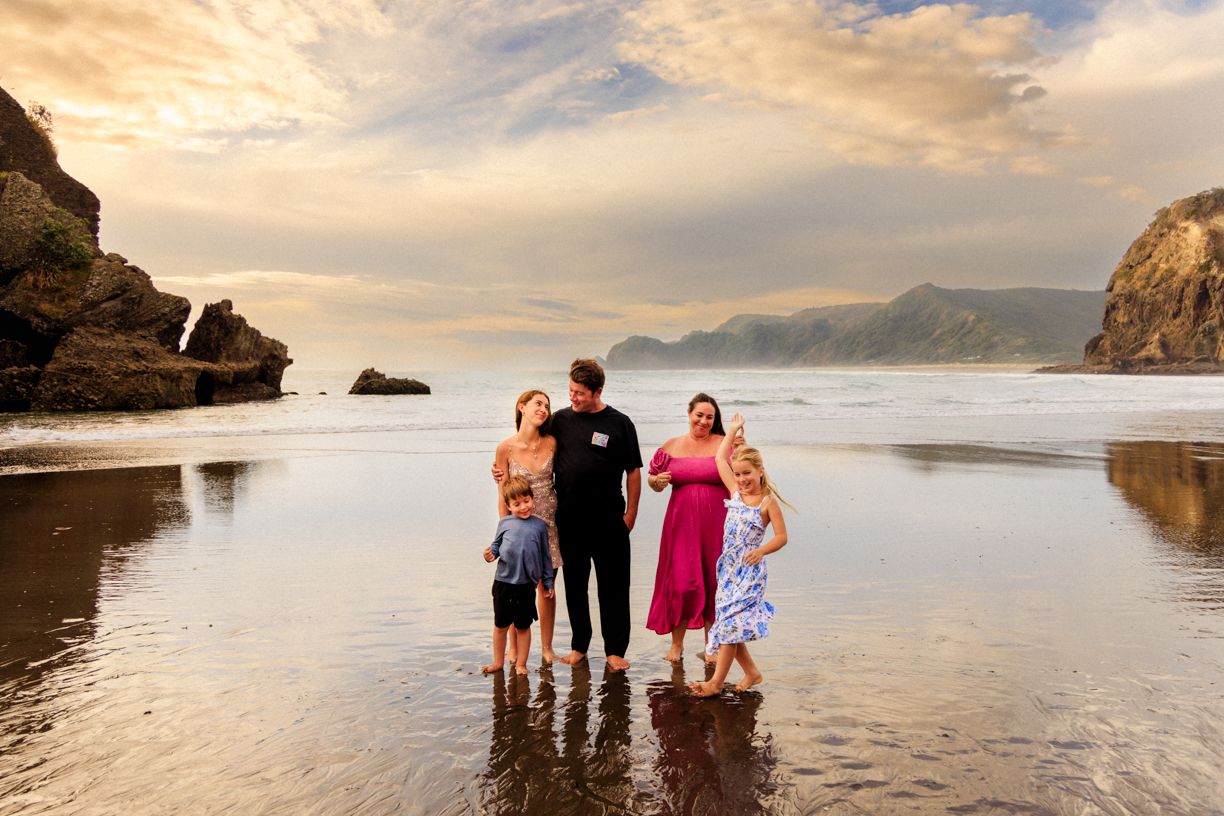 A family stands together in shallow ocean water at sunset with rocky cliffs and mountains in the background.